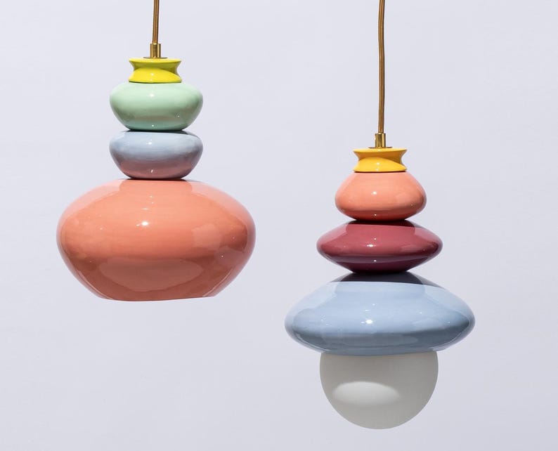 Op de afbeelding: Twee kleurrijke hanglampen met gestapelde, afgeronde vormen. E&eacute;n heeft een koraalrode basis, een lavendelkleurig middengedeelte en een mintgroene top, met een gele accent. De andere heeft een lichtblauwe basis, een bordeauxrood middengedeelte en een perzikkleurige top.