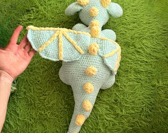 May include: A crocheted dragon toy in light blue with yellow accents. The dragon has wings, horns, and a tail. The wings and body are light blue, and the accents are yellow. The toy is soft and plush.
