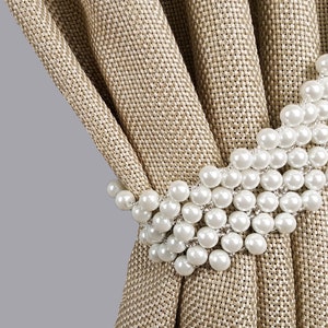 Può includere: Una tenda beige con un fermaglio per tende in perle bianche. Il fermaglio è composto da più fili di perle che sono collegati tra loro.