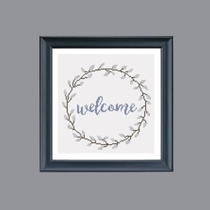May include: Cross-stitch artwork in a dark blue frame. The design features a circular wreath of gray leaves and the word "welcome" in blue script. The background is white.