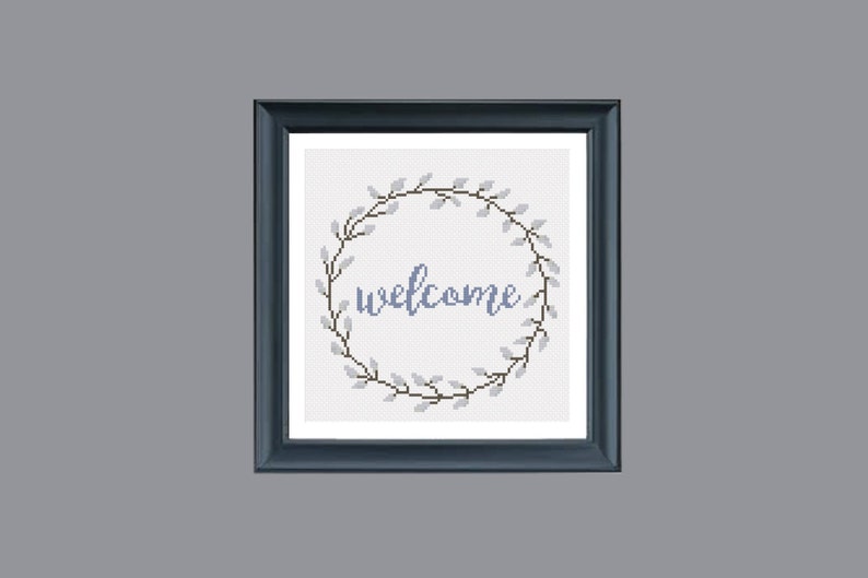 May include: Cross-stitch artwork in a dark blue frame. The design features a circular wreath of gray leaves and the word "welcome" in blue script. The background is white.