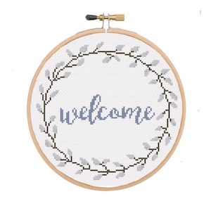 May include: A cross-stitch hoop with the word "welcome" in blue script. The word is centered on a white background and surrounded by a wreath of gray leaves and black stems. The wooden hoop is secured with a brass screw.