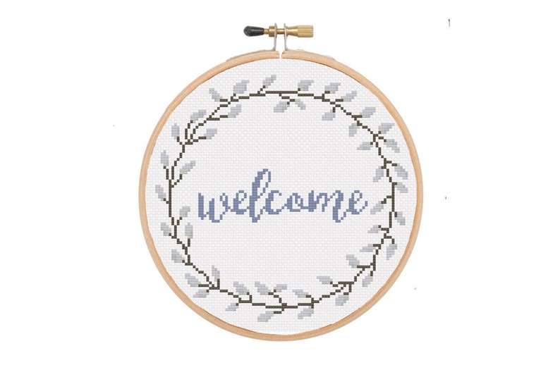 May include: A cross-stitch hoop with the word "welcome" in blue script. The word is centered on a white background and surrounded by a wreath of gray leaves and black stems. The wooden hoop is secured with a brass screw.