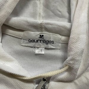 May include: Close-up of a white hooded sweatshirt with a zipper closure. The label reads "courr&egrave;ges" and the size is 38. The sweatshirt has a drawstring and a textured fabric.