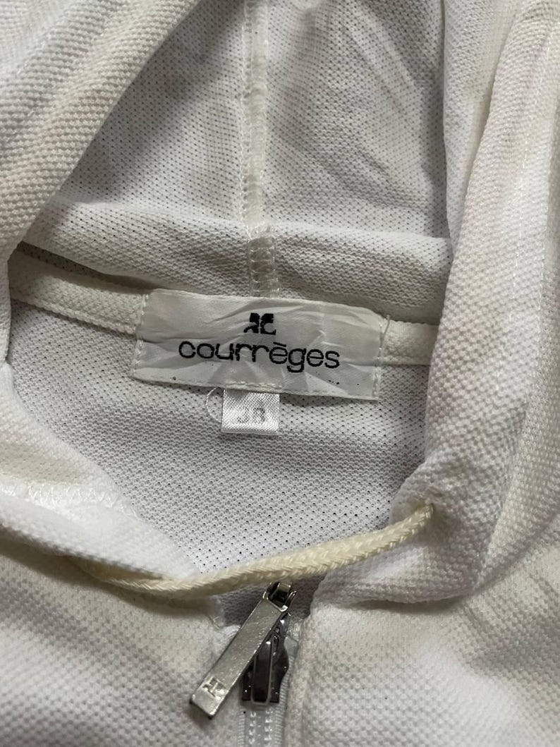 May include: Close-up of a white hooded sweatshirt with a zipper closure. The label reads "courr&egrave;ges" and the size is 38. The sweatshirt has a drawstring and a textured fabric.