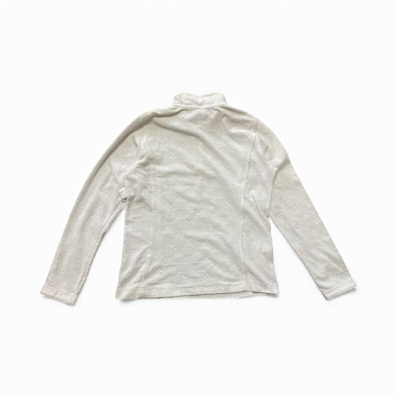 May include: A long-sleeved, off-white turtleneck shirt. The shirt has a subtle, all-over embossed pattern. The garment is made of a soft, possibly fleece-like material. The shirt is laid flat on a white surface.