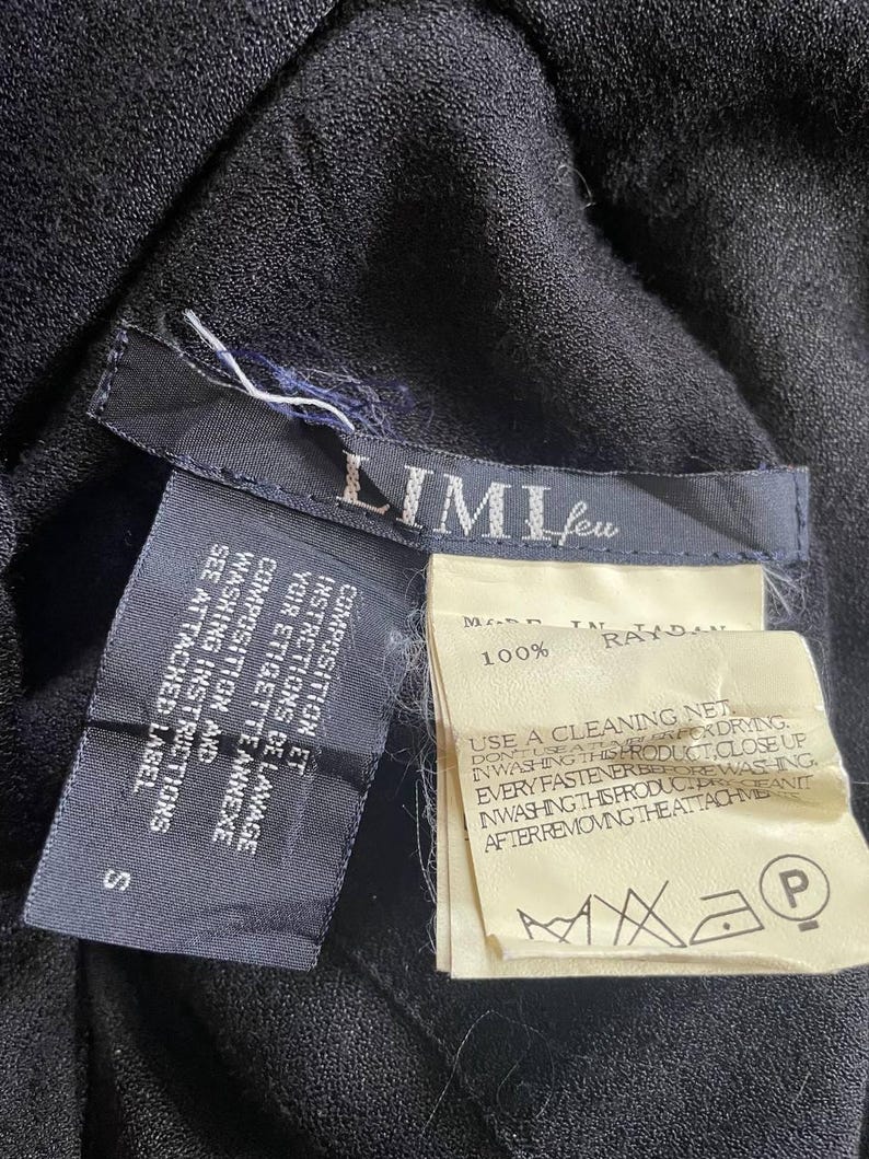 Op de afbeelding: Close-up van zwarte stof met aangehechte labels. E&eacute;n label leest "LIMI feu" en "S". Een ander label geeft aan dat de stof 100% rayon is en wasvoorschriften bevat. De labels zijn aan het kledingstuk bevestigd.