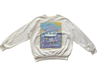 May include: Off-white sweatshirt featuring a colorful graphic of Sydney, Australia. The design includes the Sydney Harbour Bridge, sailboats, and the Sydney Opera House. The word "Sydney" is written in blue script across the top of the graphic.