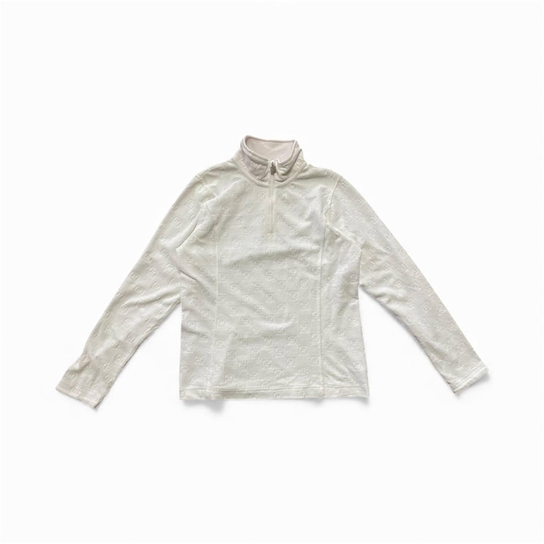 May include: A long-sleeved, off-white top with a mock neck and a quarter-length zipper. The top has a subtle, textured pattern and is laid flat on a white surface. The sleeves are long and the overall style is casual.