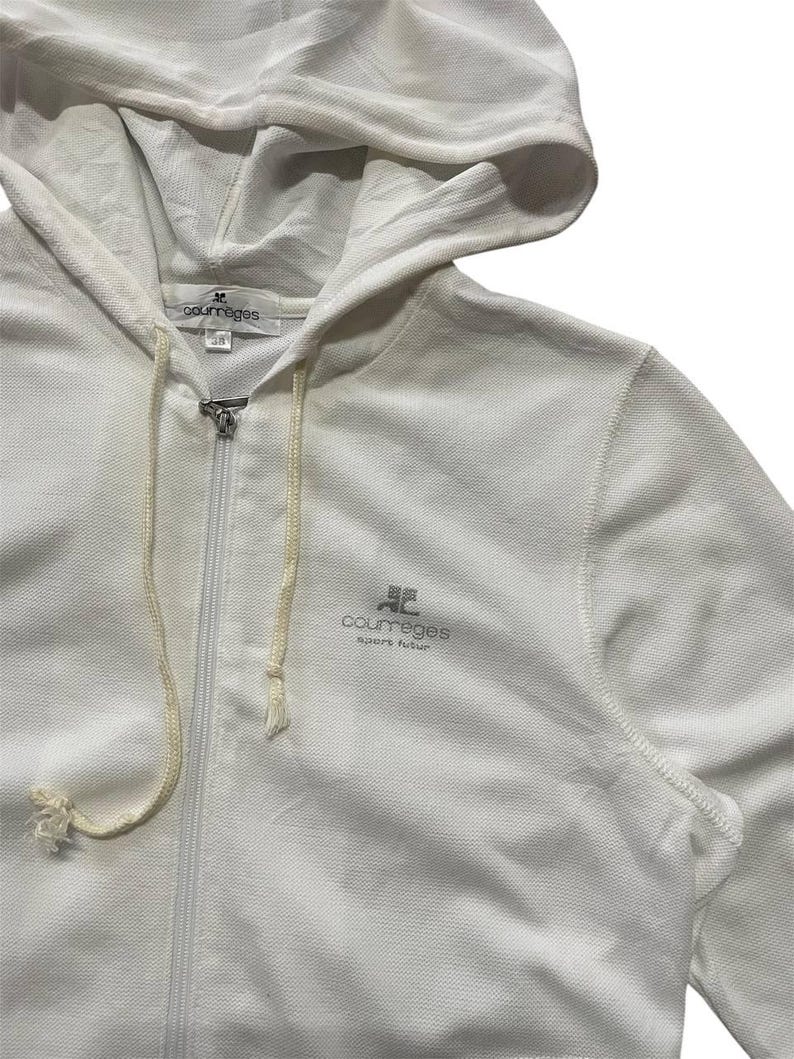 May include: A white zip-up hoodie with a drawstring hood. The hoodie features the brand name "courreges" in a gray font on the chest. The fabric appears to be a textured knit. The zipper is silver.