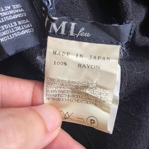 Op de afbeelding: Close-up van kledinglabels. Het hoofdlabel luidt "MI feu" en "MADE IN JAPAN 100% RAYON". Extra labels geven wasvoorschriften. De stof lijkt donker van kleur.