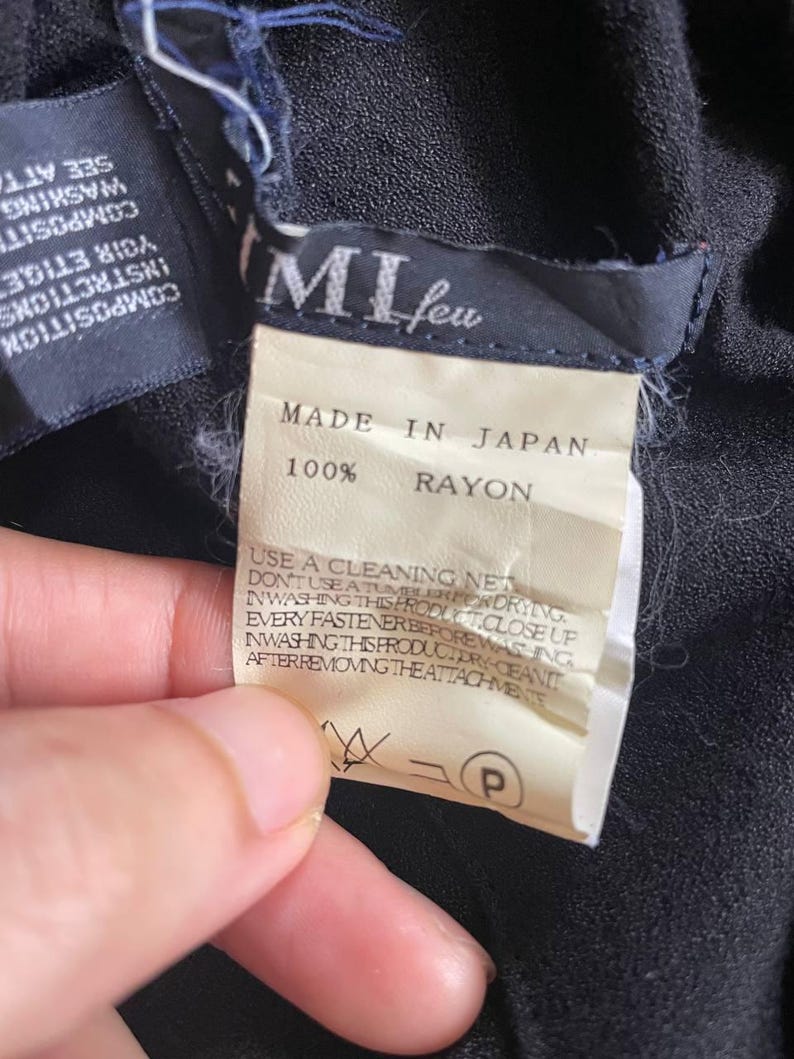 Op de afbeelding: Close-up van kledinglabels. Het hoofdlabel luidt "MI feu" en "MADE IN JAPAN 100% RAYON". Extra labels geven wasvoorschriften. De stof lijkt donker van kleur.
