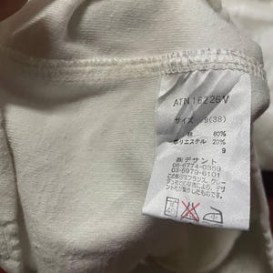 May include: Close-up of a white fabric garment tag with Japanese text and the code ATN16226V. The tag also includes washing instructions and fabric composition details. The fabric appears to be a blend of cotton and polyester.
