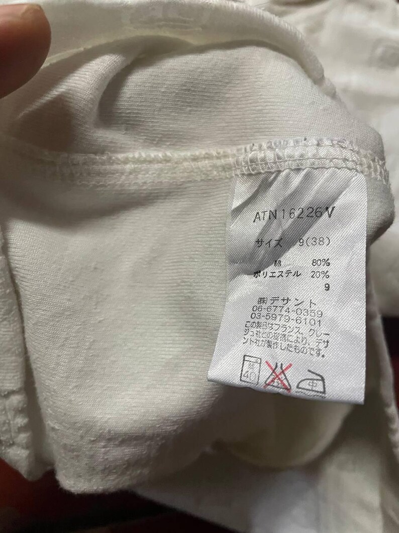 May include: Close-up of a white fabric garment tag with Japanese text and the code ATN16226V. The tag also includes washing instructions and fabric composition details. The fabric appears to be a blend of cotton and polyester.