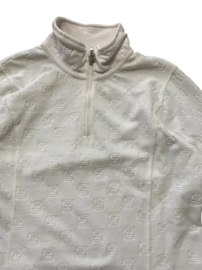 May include: White fleece pullover with a high collar and a quarter-zip closure. The fabric is patterned with a repeating embossed design. The zipper pull is silver. The garment has a soft, textured appearance.