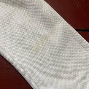 May include: Close-up of a white fabric with a textured weave. The fabric has a visible seam and some light brown stains. The background is a dark reddish-brown surface.