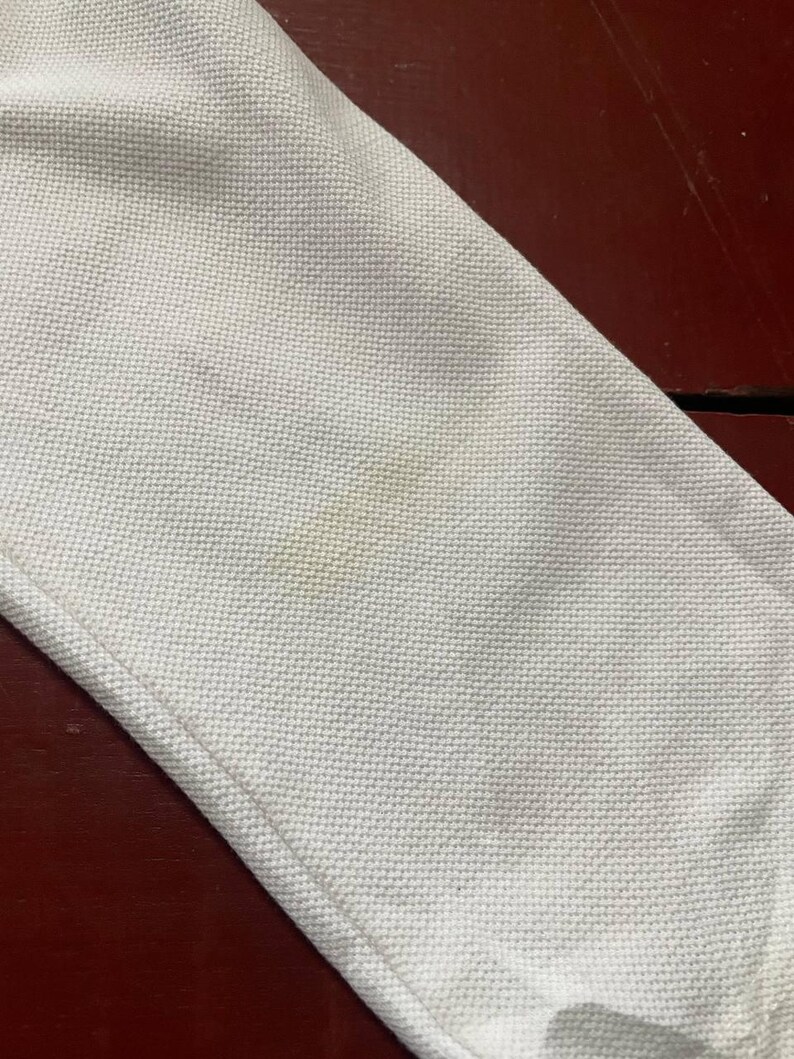 May include: Close-up of a white fabric with a textured weave. The fabric has a visible seam and some light brown stains. The background is a dark reddish-brown surface.