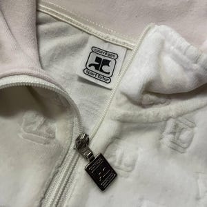 May include: Close-up of a white velour zip-up jacket. The jacket features a silver zipper and a tag with the text "courr&egrave;ges sport futur" in black. The fabric has a subtle embossed pattern.