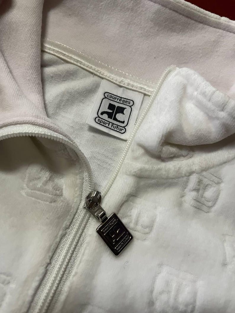 May include: Close-up of a white velour zip-up jacket. The jacket features a silver zipper and a tag with the text "courr&egrave;ges sport futur" in black. The fabric has a subtle embossed pattern.