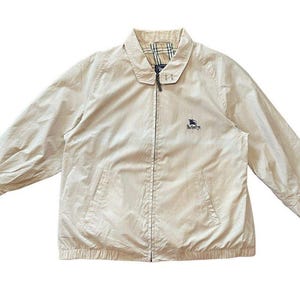 May include: A beige zip-up jacket with a collared neckline. The jacket features a small embroidered logo on the chest and a plaid pattern on the inside collar. The jacket has two front pockets and long sleeves.