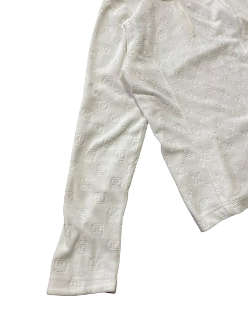 May include: White long-sleeved top with a repeating embossed pattern. The fabric appears to be soft and textured. The top has a zipper detail at the neckline and a fitted sleeve.