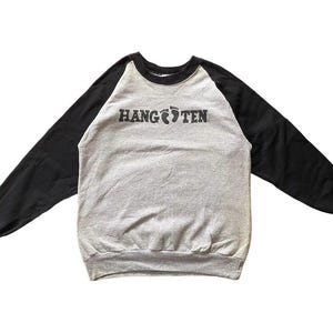 May include: A gray sweatshirt with black sleeves. The text "HANG TEN" is printed across the chest in black, with a footprint graphic between the words. The sweatshirt has a crew neck and ribbed cuffs.