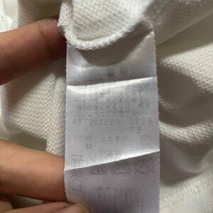 May include: A white fabric tag with text and symbols is attached to a white garment. The tag appears to be made of a smooth, satin-like material. The text is in a dark color, and the symbols are clear.