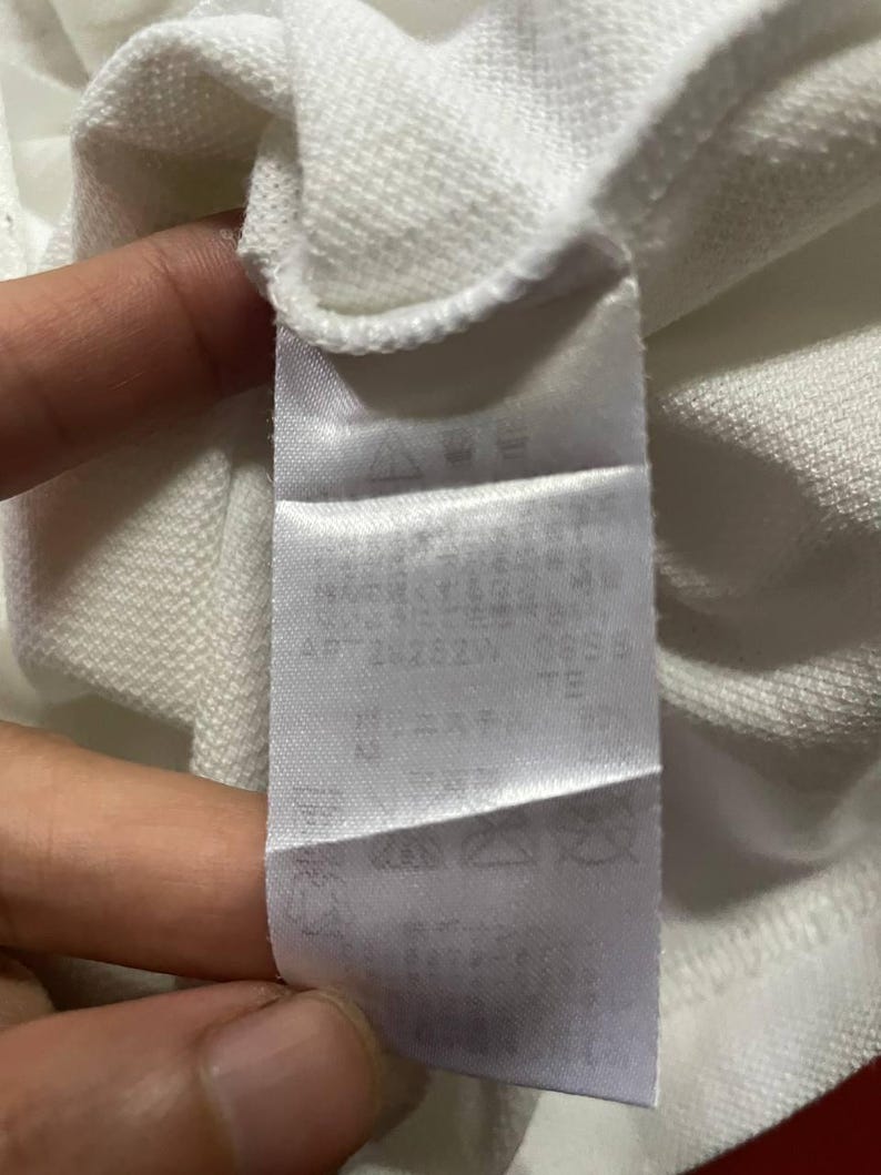 May include: A white fabric tag with text and symbols is attached to a white garment. The tag appears to be made of a smooth, satin-like material. The text is in a dark color, and the symbols are clear.
