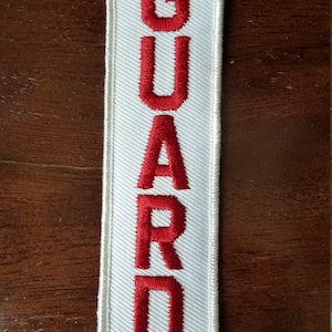 May include: White embroidered patch with the word "GUARD" in red lettering.