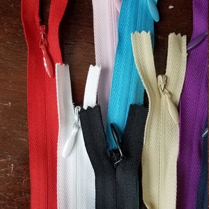 K&ouml;nnte beinhalten: Sortiment von Rei&szlig;verschlussb&auml;ndern in verschiedenen Farben, darunter Rot, Wei&szlig;, Schwarz, Beige, Lila und Blau. Die Rei&szlig;verschl&uuml;sse sind alle geschlossen und haben ein Fischgr&auml;tenmuster.