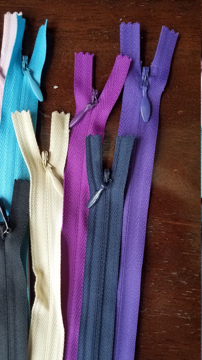 K&ouml;nnte beinhalten: Sortiment von Rei&szlig;verschlussb&auml;ndern in verschiedenen Farben, darunter Schwarz, Wei&szlig;, Lila, Blau und T&uuml;rkis. Die Rei&szlig;verschl&uuml;sse sind alle geschlossen und haben ein Fischgr&auml;tenmuster.