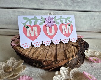 May include: A handmade greeting card with the word "MUM" in silver letters inside three pink hearts. The card is decorated with green leaves, a purple flower, and a scalloped edge. The card is displayed on a wooden slice with decorative flowers.