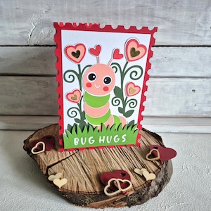 Op de afbeelding: Een handgemaakte wenskaart met een cartoonrups met een glimlach, omringd door hartvormige bloemen. De kaart heeft een rode rand en de woorden "BUG HUGS" aan de onderkant. De kaart wordt gepresenteerd op een houten schijf met hartvormige versieringen.