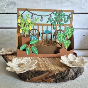 Può includere: Un diorama di carta fatto a mano con una scena di giardino in miniatura. La scena comprende piante in vaso, un tavolino con una teiera e luci a corda. Il diorama è esposto su una fetta di legno, con fiori di stoffa in primo piano.