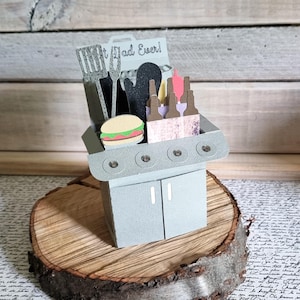 Könnte beinhalten: Ein grauer Papier-Grill mit Pfannenwender, Ofenhandschuh und Flaschen. Ein Papier-Hamburger liegt auf dem Grill. Der Text "Best Dad Ever!" befindet sich über dem Grill.
