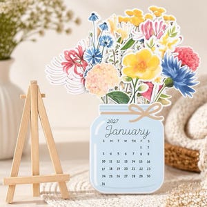 May include: A 2027 January calendar shaped like a light blue mason jar with a tan bow, featuring a colorful floral bouquet illustration on top. The calendar is displayed on a small wooden easel, showing the month and dates clearly in black text.