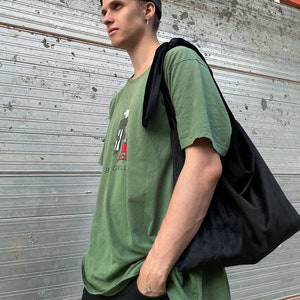 Puede incluir: Una persona lleva una camiseta verde con un gráfico en blanco y negro de una persona asando a la parrilla. La camiseta tiene el texto "Big Grill" en la parte inferior. La persona también lleva una boina negra y una bolsa de mano de terciopelo negro.