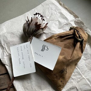May include: A white card with black text and a simple graphic design reads "You are what you eat. Stop eating little fairies, BABE." The card is on top of a brown paper bag with the word "BOSAKA" printed on it.
