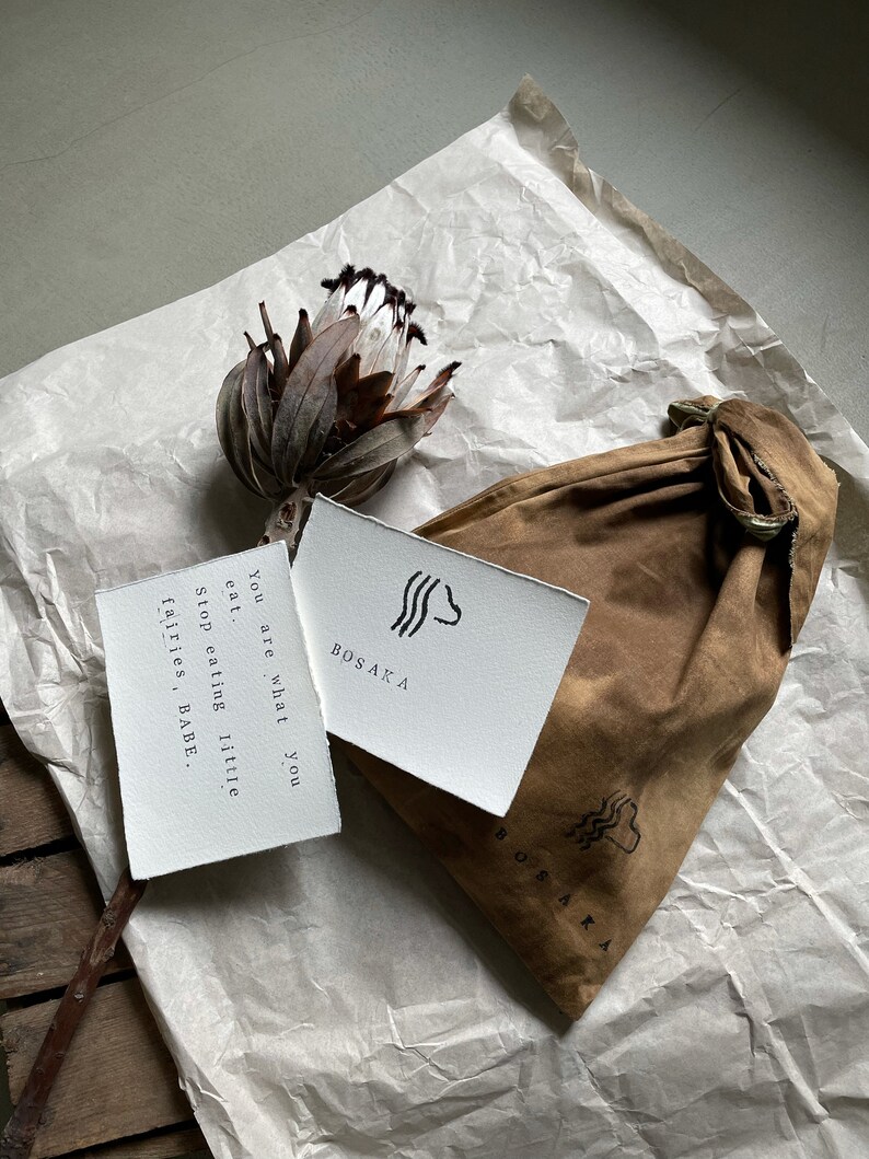May include: A white card with black text and a simple graphic design reads "You are what you eat. Stop eating little fairies, BABE." The card is on top of a brown paper bag with the word "BOSAKA" printed on it.