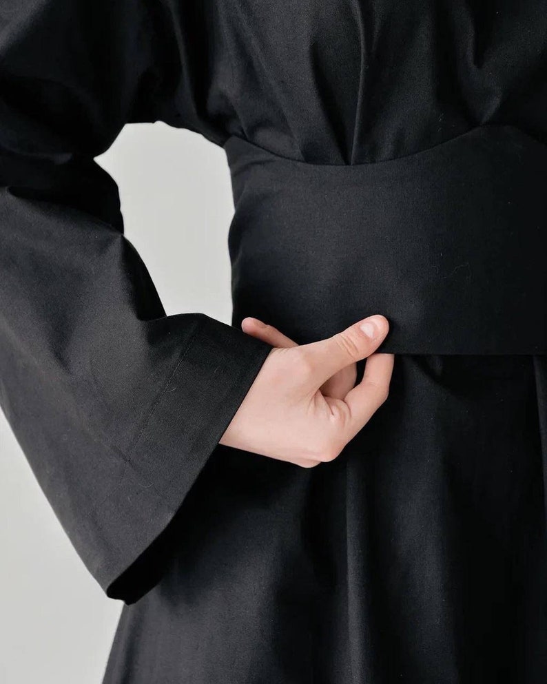 May include: A black long-sleeved dress with a wide, black belt. The dress has a loose, flowing fit and a simple, elegant design.