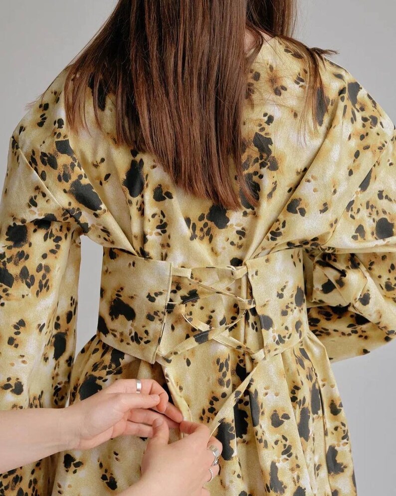 May include: A yellow and brown patterned dress with a black lace-up corset belt. The corset is being tied by two hands.