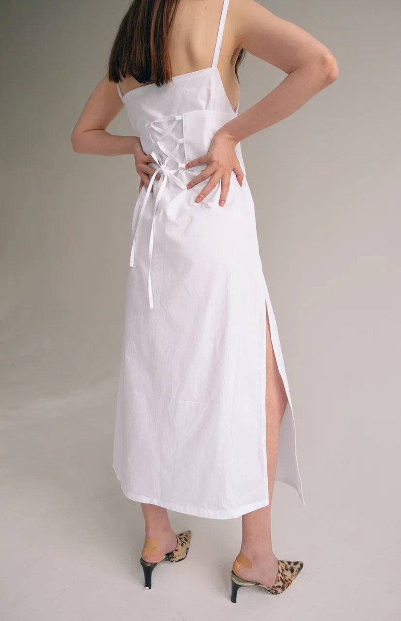 May include: A white maxi dress with a lace-up back detail and a side slit. The dress is made from a lightweight fabric and has a relaxed fit.