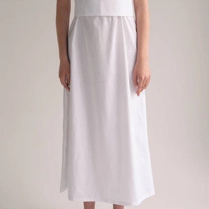 May include: A white midi dress with a fitted bodice and a flowing skirt. The dress has a square neckline and thin straps. The model is wearing the dress with leopard print heeled mules.