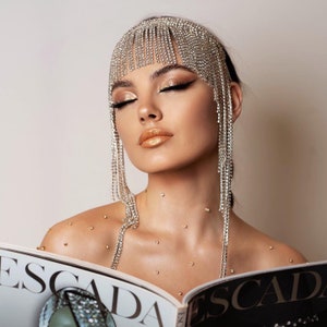 May include: A woman wearing a sparkling rhinestone headpiece with dangling strands. She is holding an open magazine with the word "ESCADA" visible. Her makeup includes gold eyeshadow and lipstick, with rhinestones on her shoulders.