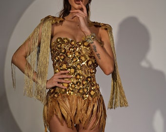 May include: A gold strapless corset top featuring dense floral sequin embellishments and long, metallic gold fringe sleeves. The garment includes a short, layered skirt section adorned with natural-toned feathers, creating a textured, shimmering appearance.