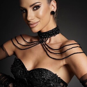 Op de afbeelding: Een vrouw draagt een zwarte strapless corset met parelversieringen en een zwarte parelketting. Ze draagt ook zwarte handschoenen en heeft haar haar in een paardenstaart gebundeld.