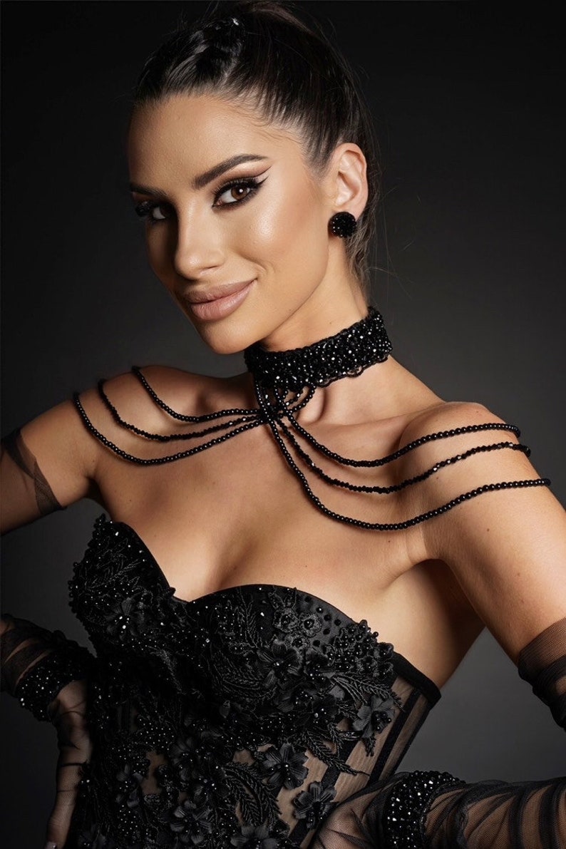 Op de afbeelding: Een vrouw draagt een zwarte strapless corset met parelversieringen en een zwarte parelketting. Ze draagt ook zwarte handschoenen en heeft haar haar in een paardenstaart gebundeld.