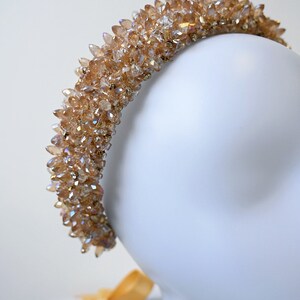 May include: A gold-colored crystal headband. The headband is covered in numerous, pointed crystals, creating a sparkling effect. The crystals are a warm, amber color. The headband is displayed on a white mannequin head.