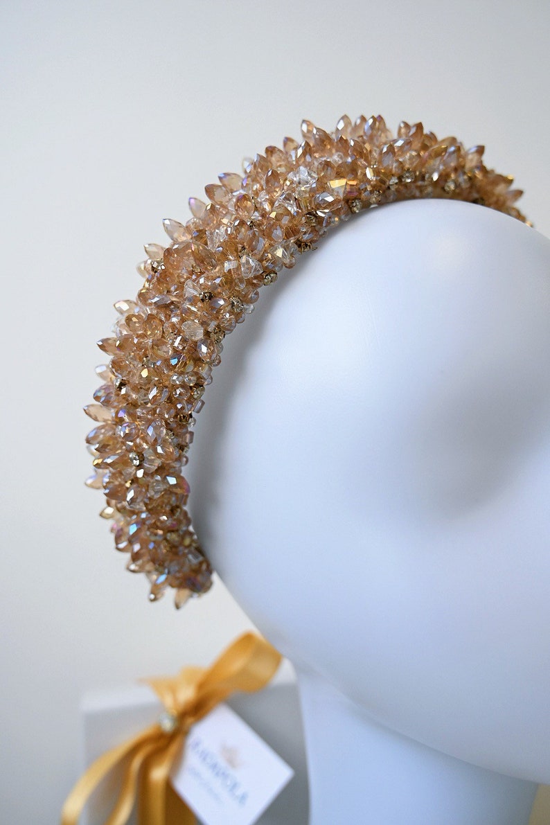 May include: A gold-colored crystal headband. The headband is covered in numerous, pointed crystals, creating a sparkling effect. The crystals are a warm, amber color. The headband is displayed on a white mannequin head.