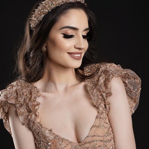 May include: A woman wearing a gold-colored, beaded tiara and a matching gown with a plunging neckline and ruffled sleeves. The dress and headpiece are adorned with sparkling embellishments, set against a dark background.